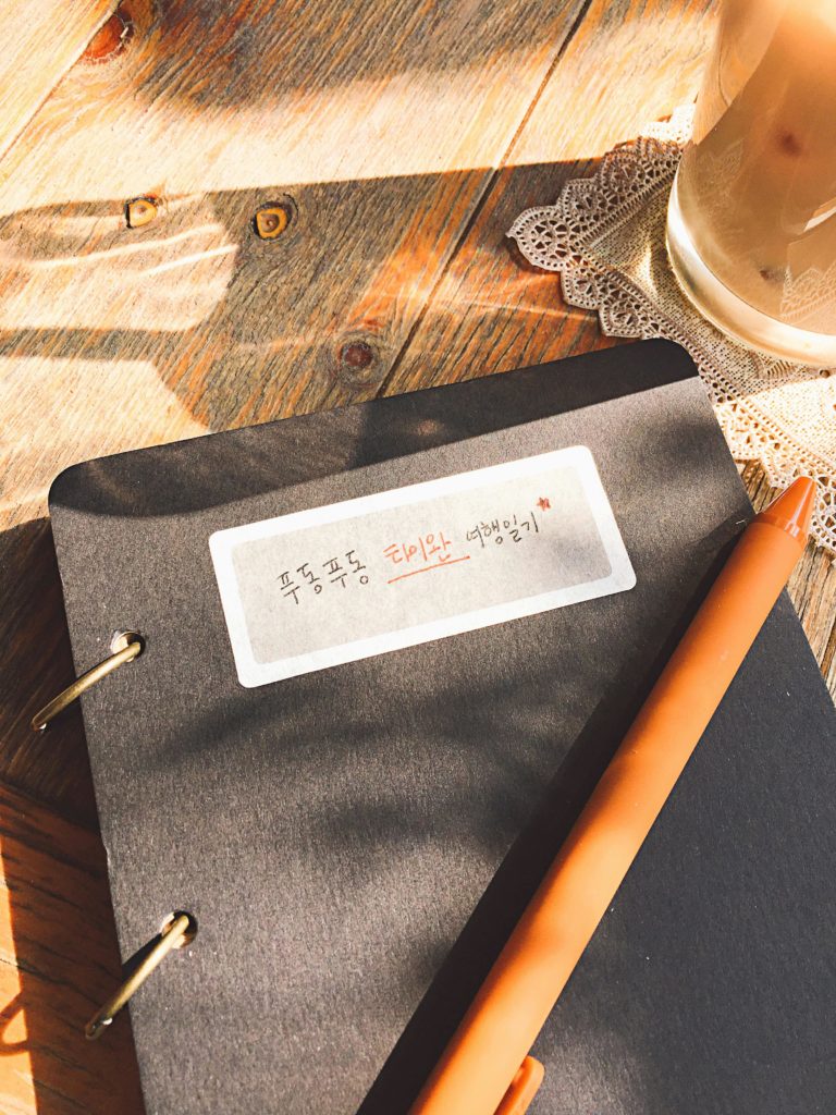 Sunlit scene of a notebook with Korean text on a wooden table, accompanied by a pen and coffee.